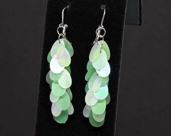 May include: A pair of dangle earrings featuring multiple overlapping, teardrop-shaped sequins in shades of light green and iridescent white. The sequins are attached to silver-toned metal hooks, creating a layered, cascading effect that hangs vertically.
