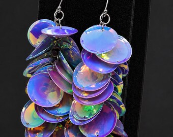 May include: A pair of dangle earrings featuring multiple iridescent, circular, purple-toned discs that reflect light with green and blue hues. The discs are layered and attached to silver-toned metal hooks, creating a cascading, reflective effect.