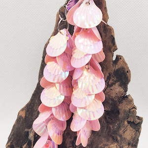 May include: A pair of pink iridescent dangle earrings with a shell-like design. The earrings are made of multiple layers of sequins and are hanging from a silver chain.