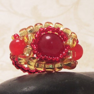 May include: A red and gold beaded ring with a large red gemstone in the center. The ring is made with small, round beads and has a delicate, feminine design.