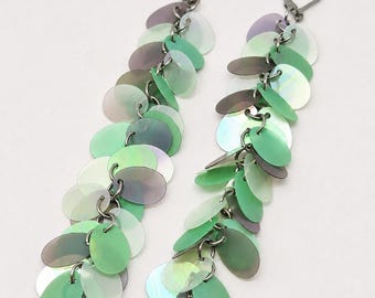 May include: A pair of long dangle earrings featuring multiple overlapping oval-shaped sequins in shades of mint green, sage, and iridescent purple. The sequins are attached to dark metal jump rings and finished with a secure lever-back closure.
