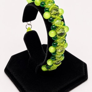 Op de afbeelding: Een kralenarmband met een dichte cluster van ronde kralen in verschillende tinten limoengroen, smaragdgroen en doorschijnend geelgroen. Het ontwerp bevat kleine zaadkralen en grotere gladde bolvormige kralen gerangschikt in een textuurpatroon.