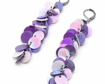 May include: A pair of long dangle earrings featuring multiple circular discs in shades of light purple, dark purple, and iridescent white. The discs are attached to dark metal lever-back closures, creating a cascading, layered, and reflective effect.