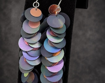 May include: A pair of long dangle earrings featuring multiple overlapping circular discs in iridescent shades of dark blue, purple, and copper. The discs are suspended from silver-toned metal hooks, creating a layered, cascading effect with a metallic finish.