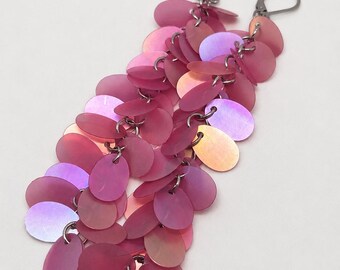 May include: A pair of dangle earrings featuring multiple overlapping oval-shaped pink iridescent sequins attached to dark metal jump rings and secured with dark metal lever-back closures. The sequins display a shifting color effect from pink to orange.