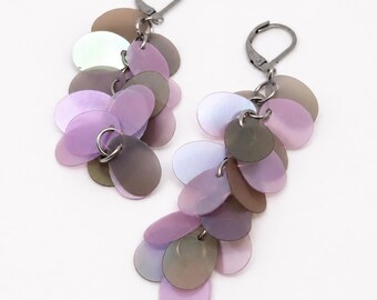 May include: A pair of dangle earrings featuring multiple overlapping circular discs in shades of iridescent lavender, soft purple, and muted grey. The discs are attached to dark gunmetal-toned leverback closures, creating a cascading cluster effect.