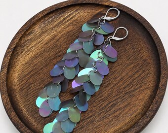 May include: A pair of long dangle earrings featuring multiple iridescent, oval-shaped discs in shades of purple, blue, and green. The discs are linked together in a cascading pattern and attached to silver-toned metal lever-back earring hooks.