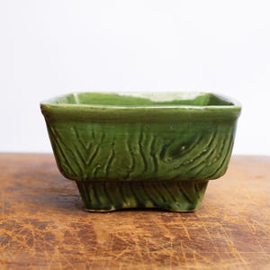 Puede incluir: Una jardinera cuadrada de cerámica verde con un diseño texturizado de vetas de madera. La jardinera tiene una base elevada y está colocada sobre una superficie de madera. La jardinera mide aproximadamente 15 cm de ancho y 10 cm de alto.
