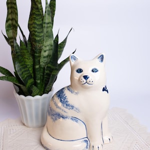 May include: A white ceramic cat ornament with blue detailing, including eyes, stripes, and a bow. It sits on a cream lace doily, with a potted snake plant to the left. The cat has blue markings.