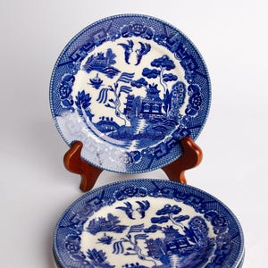 May include: A set of round ceramic plates featuring a blue and white willow pattern. The design includes detailed illustrations of pagodas, willow trees, birds, and a bridge. One plate is displayed upright on a wooden stand, while two others are stacked.