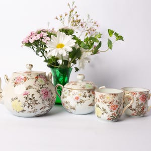Puede incluir: Un juego de té vintage con estampados florales, que incluye una tetera, un azucarero y dos tazas. El juego es de color crema con diseños florales rosas, amarillos y marrones. Un jarrón verde con flores está en el fondo.