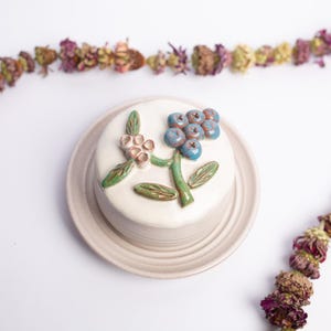 May include: A ceramic butter dish with a white glaze, decorated with a green stem, leaves, and blue berries. The dish sits on a matching plate, and is surrounded by a garland of dried flowers. The dish is approximately 4 inches in diameter.