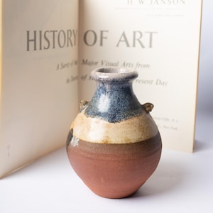 May include: A small, handcrafted ceramic vase with a rounded body and a narrow neck. The vase features a color palette of brown, beige, and blue, with small handles on either side. The vase is set in front of an open book titled "History of Art."