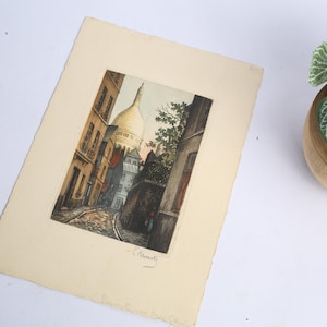May include: A rectangular paper print featuring a colored etching of a narrow cobblestone street in Paris leading toward the Sacré-Cœur Basilica. The artwork is signed in the bottom right corner and is mounted on a cream-colored paper border.