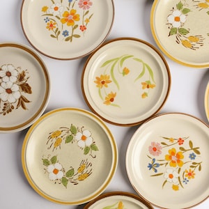 May include: A collection of round ceramic plates featuring various floral patterns in shades of orange, yellow, brown, and green. Each plate has a distinct botanical design, including clusters of daisies, wheat-like stalks, and leafy stems.