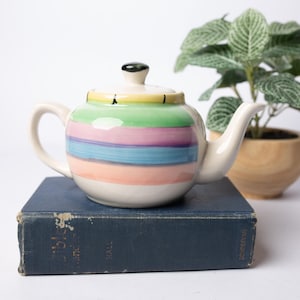 Puede incluir: Una tetera de cerámica color crema con rayas horizontales en colores pastel. La tetera está colocada sobre un libro azul vintage, con una pequeña planta en una maceta de madera en el fondo.