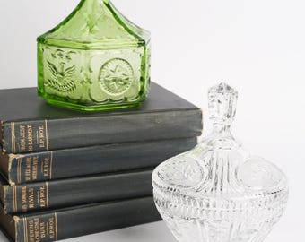 May include: A hexagonal green glass candy dish with a pointed lid featuring embossed eagle and star motifs, alongside a clear glass footed candy dish with a faceted lid and intricate pressed patterns, both resting near a stack of four black books.