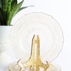 May include: A clear, decorative glass plate with a scalloped edge and intricate floral design, displayed on a gold-colored stand. A smaller matching plate sits below. The plate is approximately 10 inches in diameter.