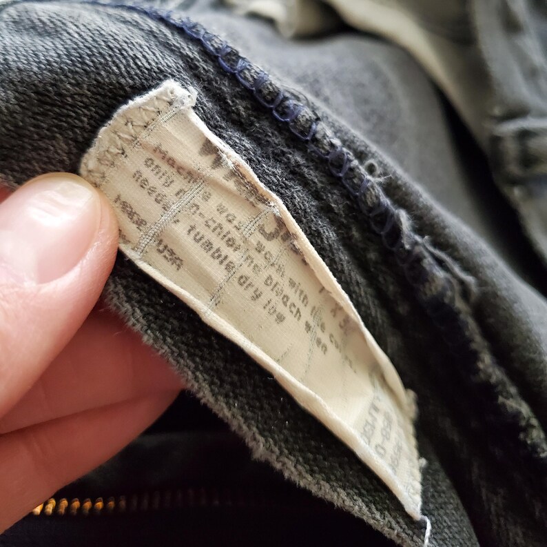 May include: A white care label sewn into a black garment. The label has instructions for washing and drying the garment. The text on the label reads "Machine wash with like colors, non-chlorine bleach when needed, tumble dry low, Made in USA."