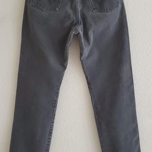May include: A pair of dark gray denim jeans with a straight leg fit. The jeans have a classic five-pocket design and a leather patch on the back waistband with the Wrangler logo.