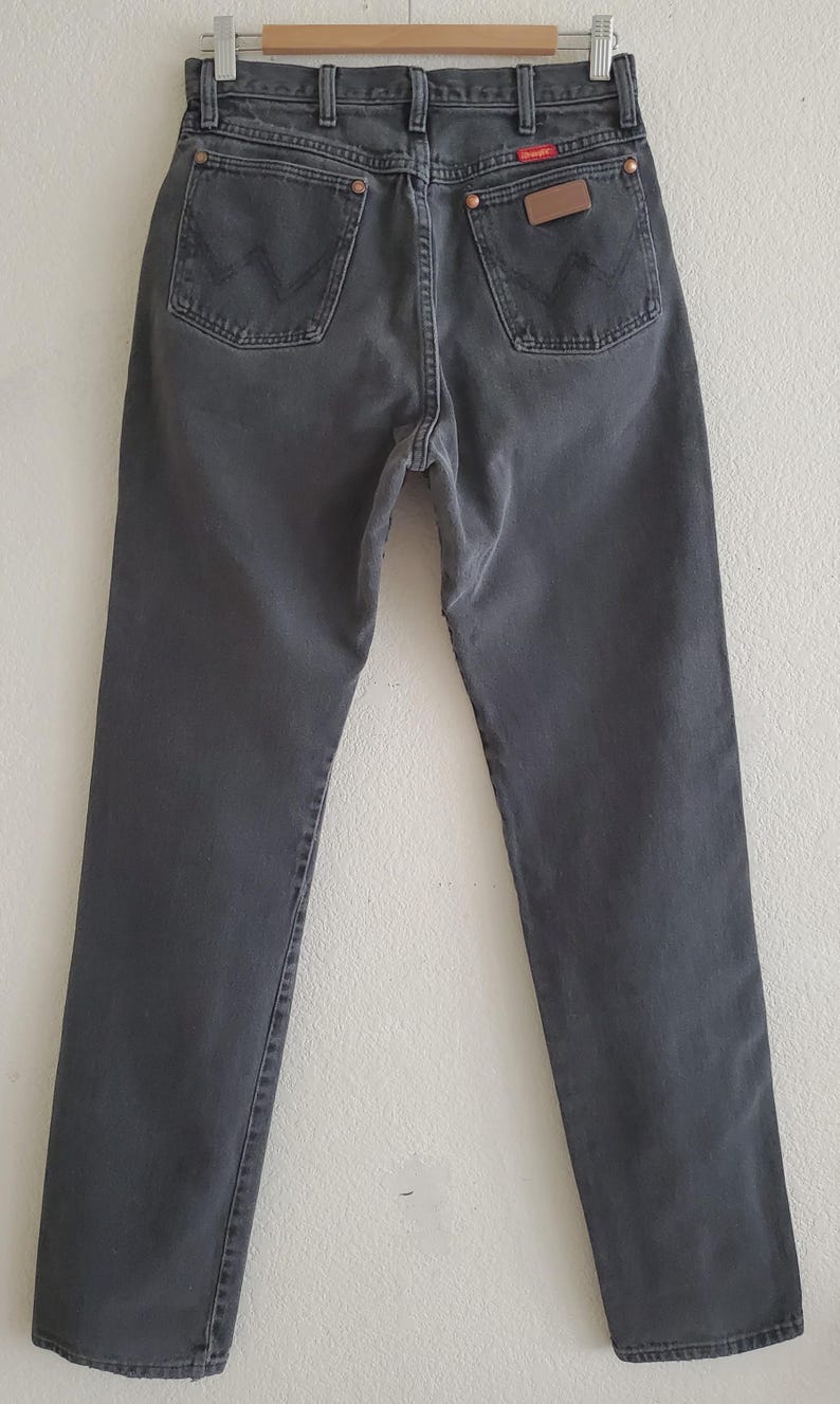 May include: A pair of dark gray denim jeans with a straight leg fit. The jeans have a classic five-pocket design and a leather patch on the back waistband with the Wrangler logo.