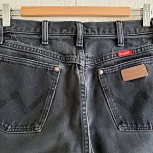 May include: Black denim jeans with a brown leather patch on the back pocket and a red tab with the word "Wrangler" on the right back pocket.
