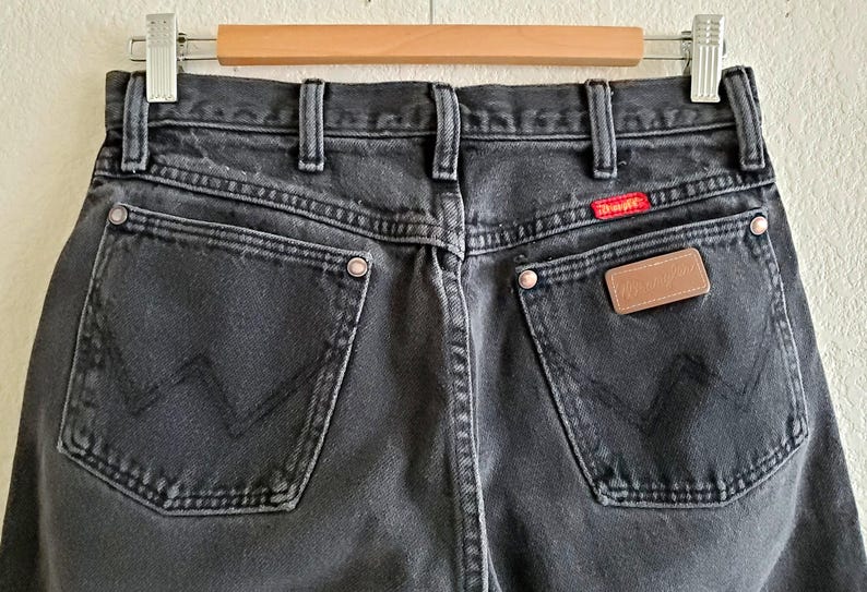 May include: Black denim jeans with a brown leather patch on the back pocket and a red tab with the word "Wrangler" on the right back pocket.