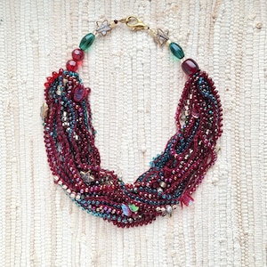 May include: A statement necklace with multiple strands of burgundy, teal, and iridescent beads. The necklace has a gold-tone clasp and is adorned with star-shaped charms and green and red beads. The design creates a bohemian style.