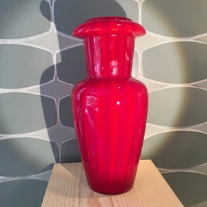 May include: A red glass vase with a wide mouth and a ribbed design. The vase is sitting on a wooden surface.