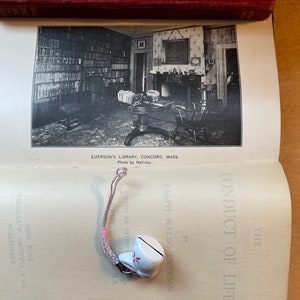 Może przedstawiać: Otwarta książka z czarno-białym zdjęciem wnętrza biblioteki. Tytuł książki to "The Conduct of Life" autorstwa Ralpha Waldo Emersona. Do książki przymocowany jest mały biały dzwonek z r&oacute;żową wstążką.