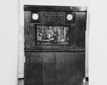 May include: A black and white photo of a wooden cabinet with two round windows and a painting of three people inside. The cabinet has a sign that reads "Welgelegen en hac spijt".