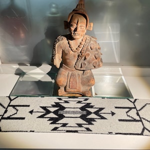 Puede incluir: Una estatua de cerámica marrón de una figura Maya que lleva un tocado y un collar de cuentas. La estatua está sentada sobre una superficie de vidrio con una alfombra de patrón blanco y negro delante de ella.