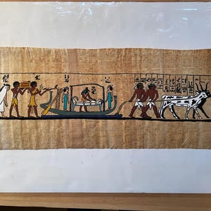 May include: A hand-painted papyrus scroll depicting an ancient Egyptian procession. The scene shows figures carrying a sarcophagus on a boat, followed by people pulling a cart with a cow. The scroll is decorated with hieroglyphs and has a beige background.