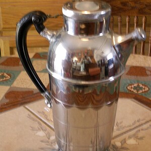 May include: A vintage chrome cocktail shaker with a black handle and a spout. The shaker has a cylindrical body with horizontal lines and a rounded top. The chrome finish reflects the surrounding environment.