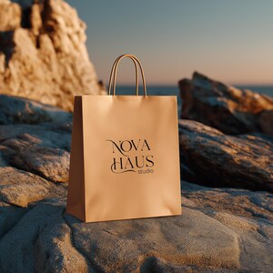 May include: A tan paper shopping bag with the text "NOVA HAUS studio" in black script. The bag has matching tan handles and is sitting on a gray rock. The background includes a rocky cliff and the ocean.