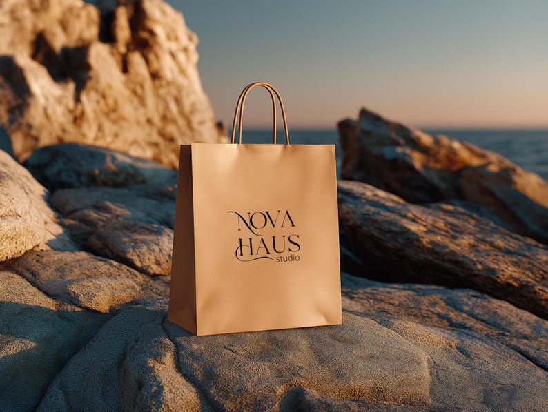 May include: A tan paper shopping bag with the text "NOVA HAUS studio" in black script. The bag has matching tan handles and is sitting on a gray rock. The background includes a rocky cliff and the ocean.