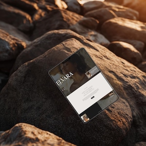May include: A tablet displaying a website with the text "ELVARA" on the screen. The tablet is resting on a large, dark rock, with other rocks and the ocean in the background. The website features images and text.