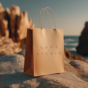 May include: A tan shopping bag with the text "NOVA STUDIO" in a minimalist font. The bag has thin, beige handles and is set against a rocky, coastal backdrop with the ocean visible in the distance.