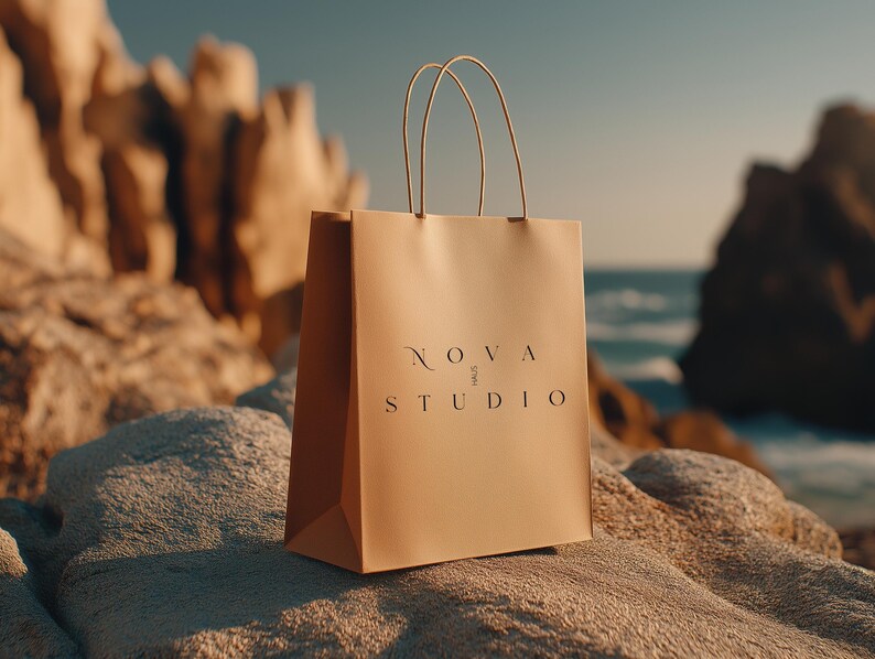 May include: A tan shopping bag with the text "NOVA STUDIO" in a minimalist font. The bag has thin, beige handles and is set against a rocky, coastal backdrop with the ocean visible in the distance.