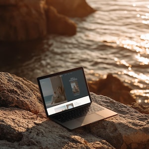 May include: A laptop computer with a black frame and screen sits open on a large, textured rock near the ocean. The screen displays a website with the text "SOLA COACHING" and images of people. The scene is bathed in warm sunlight.