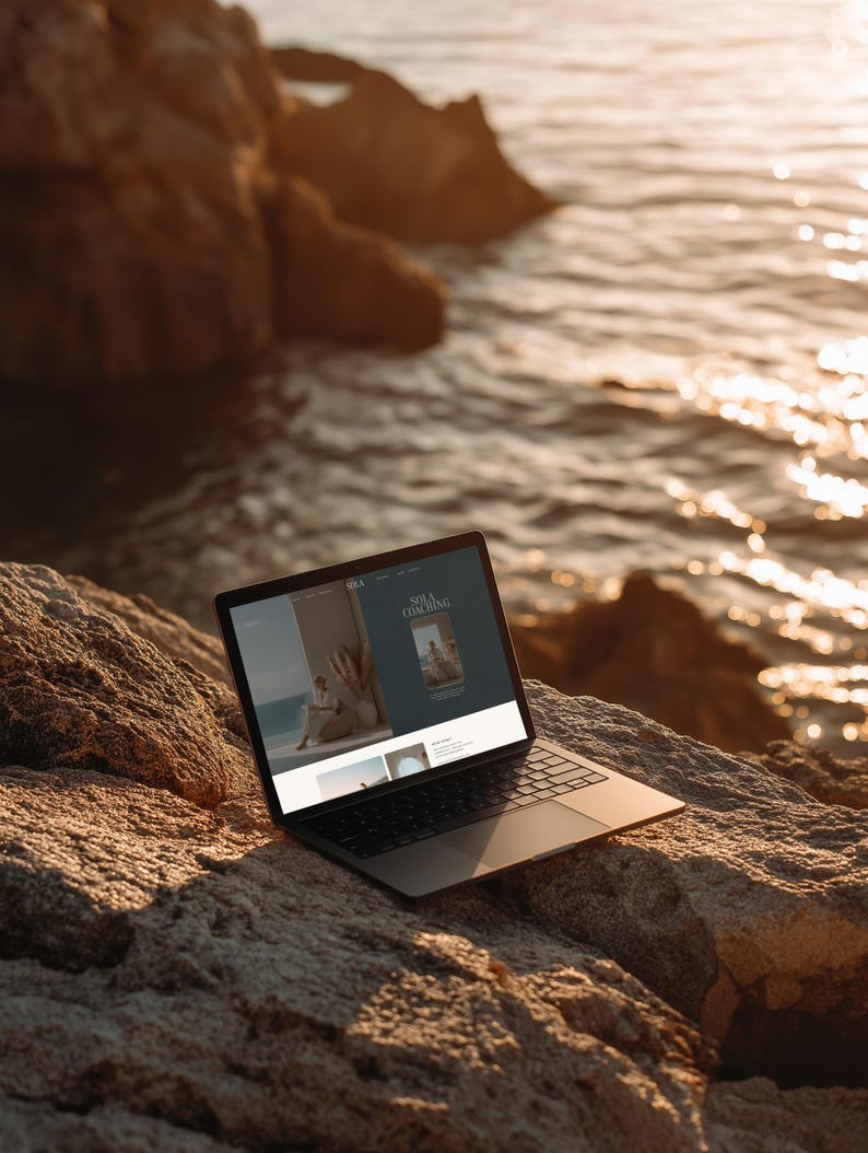 May include: A laptop computer with a black frame and screen sits open on a large, textured rock near the ocean. The screen displays a website with the text "SOLA COACHING" and images of people. The scene is bathed in warm sunlight.