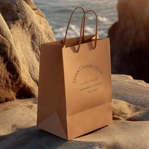 May include: A tan paper shopping bag with brown handles, resting on a sandy surface near the ocean. The bag features the text "EMMA CHARLOTTE worldwide traveler BLOGGER & AUTHOR EST. 2014" in a circular design.