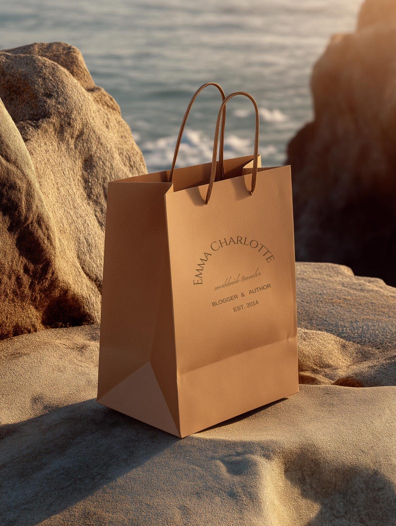 May include: A tan paper shopping bag with brown handles, resting on a sandy surface near the ocean. The bag features the text "EMMA CHARLOTTE worldwide traveler BLOGGER & AUTHOR EST. 2014" in a circular design.