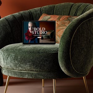 Może przedstawiać: Ciemnozielony fotel z aksamitu ze złotymi nogami. Na krześle znajduje się tablet z napisem "BOLD STUDIO". Za tabletem znajduje się dekoracyjna poduszka z pomarańczowo-zielonym wzorem liści.