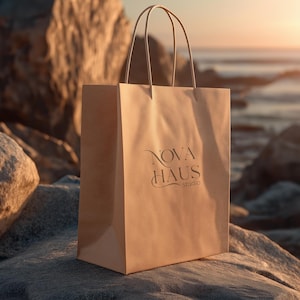 May include: A tan paper shopping bag with the text "NOVA HAUS studio" printed on the front. The bag has a rectangular shape with a flat bottom and two handles. The bag is sitting on a rock with a beach and sunset in the background.