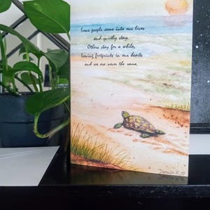 May include: Watercolor art print featuring a sea turtle on a sandy beach under a setting sun. The print includes handwritten text: "Some people come into our lives and quietly stay. Others stay for a while, leaving footprints in our hearts and we are never the same."