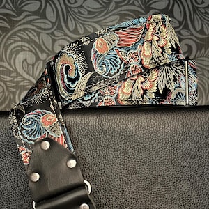 Könnte beinhalten: Ein schwarzer Gitarrengurt mit einem farbenfrohen Paisley-Muster in Rot, Blau und Gold. Der Gurt hat ein schwarzes Lederendstück mit silbernen Nieten und einem dekorativen Emblem.