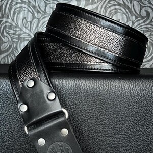 May include: Black leather guitar strap with a textured silver center stripe. The strap features a black leather end piece with silver rivets and a circular embossed logo. The strap is shown on a black textured surface.