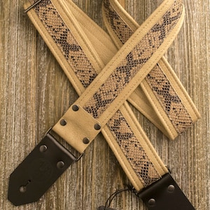 May include: A tan leather guitar strap with a black snake skin pattern. The strap has black leather ends with metal buckles and a Hang Over brand tag.