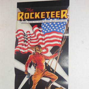 May include: A vintage poster advertising the Super Nintendo Entertainment System (SNES) video game "The Rocketeer". The poster features a red, white, and blue American flag with a cartoon image of the Rocketeer in his red and gold suit, holding a gun and standing on a building.